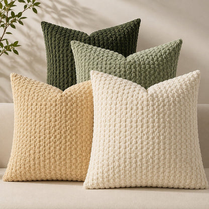 Quatre housses de coussin Cocon Naturel vert foncé, vert sauge, beige et écru sur un canapé, avec texture bouclée.